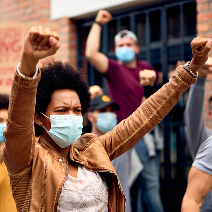 African-American-woman-wearing-protective-face-mask-while-protesting-with-arms-raised-on-city-streets